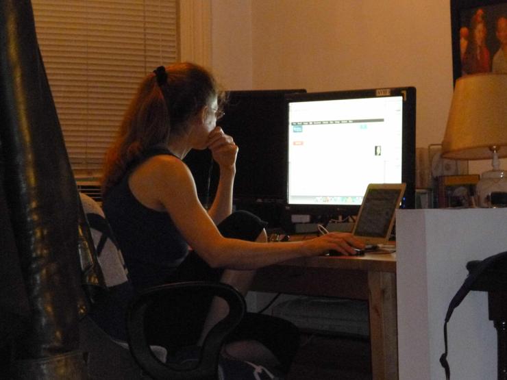 Color photo of Sabina Knight working at a wooden desk facing a large monitor. A small laptop sits in front and to the side of the desktop monitor.

Sabina is seen from the three-quarter right back view (i.e., the viewer's left).

Her long strawberry blond hair is tied back in a ponytail with a black scrunchie.

The shades of the window to her left are down. So the only lighting is artificial. Except for the computer monitor, the lighting is minimal. The dimness suggests that the photo was taken after dark.